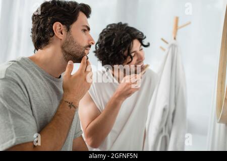 tattooed man touching face while boyfriend brushing teeth on blurred background Stock Photo