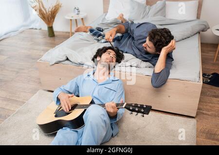 gay man playing acoustic guitar near caring boyfriend on bed Stock Photo