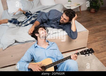 smiling gay man playing acoustic guitar near bearded boyfriend on bed Stock Photo