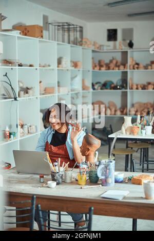 Joyful female ceramist using laptop in pottery workshop Stock Photo
