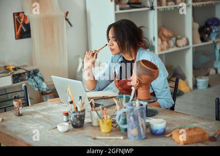 Beautiful young woman potter using laptop in pottery workshop Stock Photo