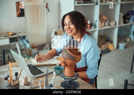 Cheerful woman ceramist using laptop and writing on notebook Stock Photo