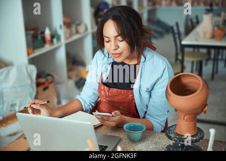Charming female potter using laptop and smartphone in workshop Stock Photo