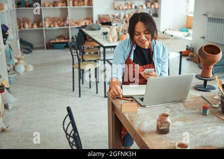 Joyful woman using laptop and smartphone in pottery workshop Stock Photo