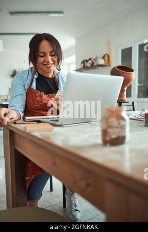 Cheerful woman using smartphone and laptop in pottery workshop Stock Photo