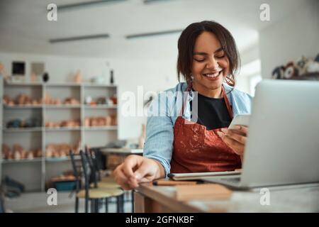 Cheerful woman using mobile phone and laptop in pottery workshop Stock Photo