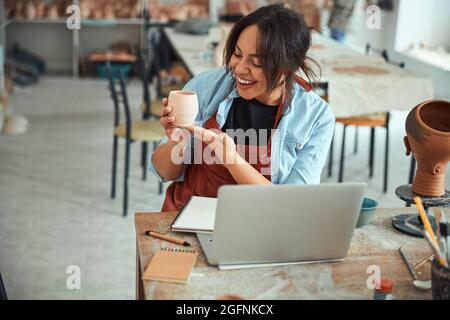 Cheerful female potter holding clay pot and using laptop Stock Photo
