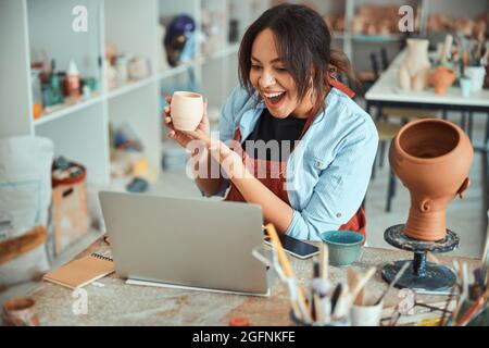 Joyful female potter holding clay pot and using laptop in workshop Stock Photo