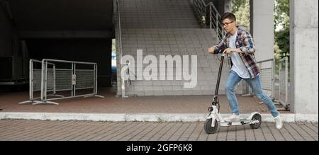 A young businessman in a shirt and glasses starts riding an electric scooter. The modern entrepreneur uses modern ecological transport Stock Photo
