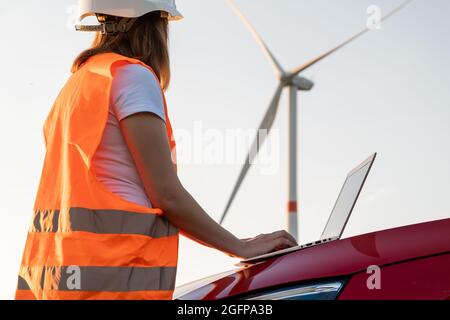 An windmill engineer supervises the work of the wind turbines on the laptop standing near the car. Generation of eco green electricity.  Stock Photo