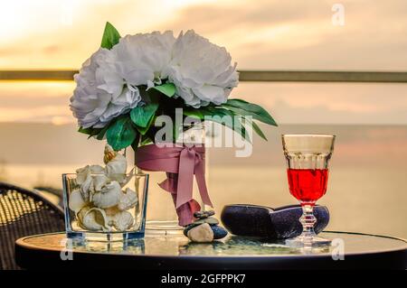 two glasses of wine against the backdrop of the sunset by the sea, on a table on the balcony, seashells and flowers Stock Photo