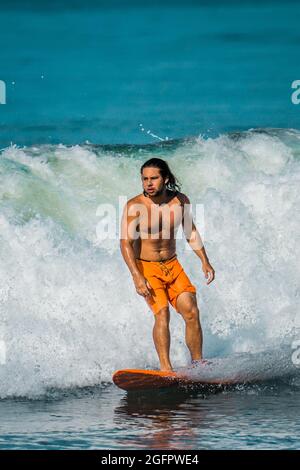 Playa Hermosa, Guanacaste, Costa Rica - 07.26.2020: A muscular long-haired latino man wearing orange shorts is surfing at the pacific Coast of Costa R