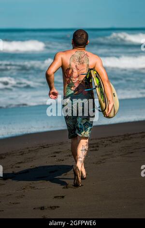 Playa Hermosa, Guanacaste, Costa Rica - 07.26.2020: A well built man with colorful tattoos is carrying a surfboard and running towards the ocean.
