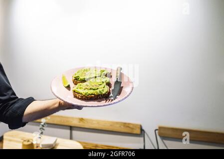 Male hand holding the pink plate with two sandwiches with avocado and sesame seeds and cutlery Stock Photo