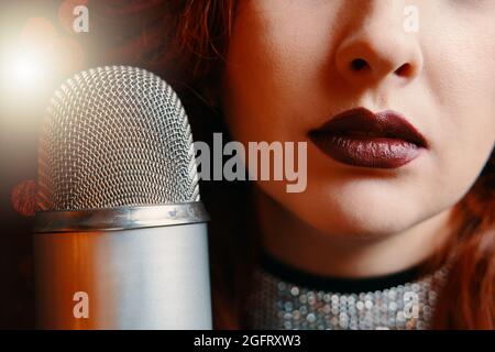 Close-up of women's lips painted with burgundy lipstick and retro microphone. Female singer with disco mic on bokeh light background. Stock Photo