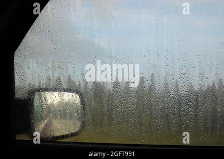 Raindrops on a vehicle window, looking out at rear view mirror, forest and cloudy sky during a heavy rain storm Stock Photo