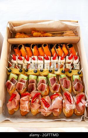 Snack food in cardboard box with small sandwiches, eclairs, bruschetta with cold cuts, cheese and seafood for catering buffet for party. Stock Photo