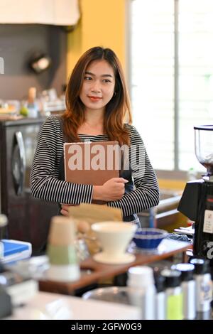 Portrait of cheerful female barista smiling, looking away, standing near coffee machine at coffee shop Stock Photo
