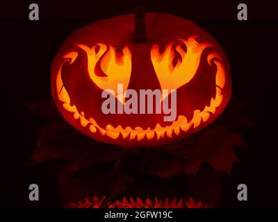 Illuminated, smiling, large, hand made pumpkin with eyes like fire flames on maple leaves in the dark halloween night Stock Photo