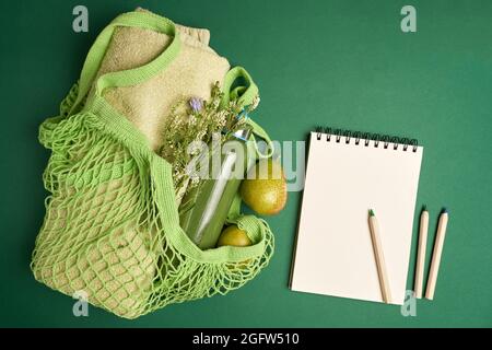 Green string bag with fruits, towel, bottle, paper notepad and pencils on green Stock Photo