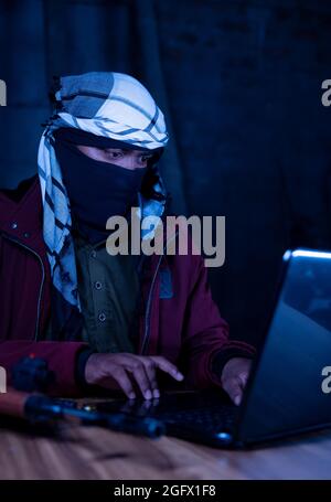 Islamic militant with face cover and gun on table with laptop to hack - concept of modern cyber terrorism or cyber attacks Stock Photo