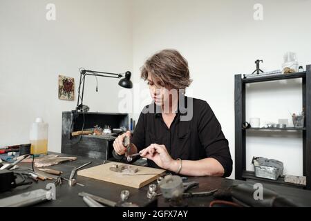 Busy middle-aged woman in black shirt sitting at desk in workshop and soldering Jewelry while finishing work Stock Photo