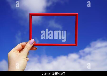 Red rectangular horizontal frame in female hand against blue sky. Sunny day background. Bright space for your text Stock Photo