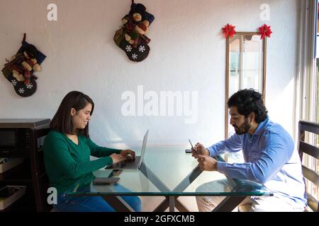 Cheerful young Hispanic couple or friends sitting around a table using their devices Stock Photo