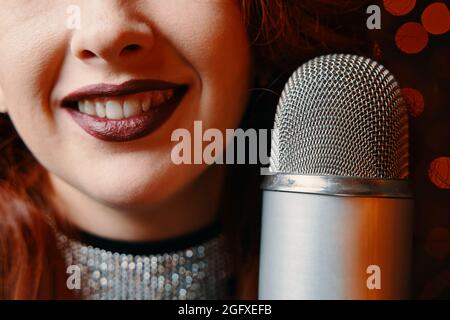 Close-up of smiling female singer's and retro microphone on bokeh blur background. Red-haired cheerful woman with burgundy lipstick on her lips perform at concert. Night party in club or restaurant. Stock Photo