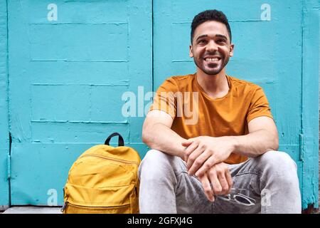 Happy handsome young Latino man sitting with a backpack on the side. Taking a rest from a walk in the city. . High quality photo Stock Photo