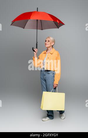 senior woman in jacket and jeans standing with shopping bag and smiling under umbrella on grey Stock Photo