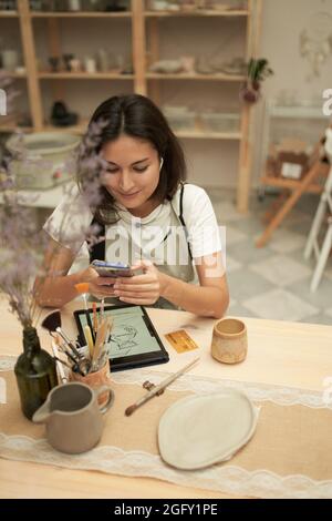 High angle of female artisan smiling and using smartphone while sitting at table with tablet and crockery in professional workshop Stock Photo