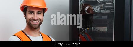Smiling workman in hard hat looking at camera near switchboard, banner Stock Photo