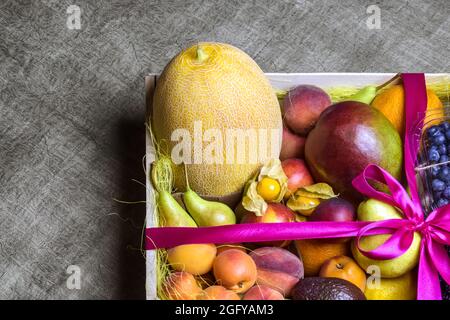 Juicy ripe fruits and berries are beautifully stacked in wooden box tied with pink ribbon. Dark background. Food delivery, gift fruit set.. Copy space Stock Photo