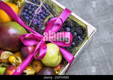 Juicy ripe fruits and berries are beautifully stacked in wooden box tied with pink ribbon. Dark background. Food delivery, gift fruit set. Copy space Stock Photo