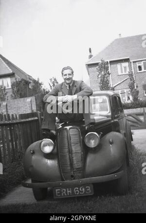 1950, historical, outside in a side area of a suburban housing area, a man in a wool suit posing for a photo, sitting on the bonnet of a motorcar of the era, England, UK. Stock Photo