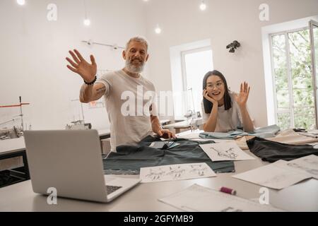 Smiling interracial designers waving hands at laptop near sketches and fabric Stock Photo