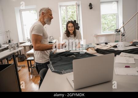 Smiling multiethnic designers looking at sketch near fabric and laptop Stock Photo