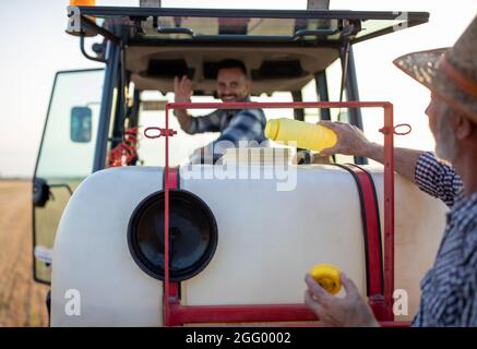 Senior farmer pouring chemicals in sprayer for crop protection in field. Younger man sitting in tractor and looking back Stock Photo