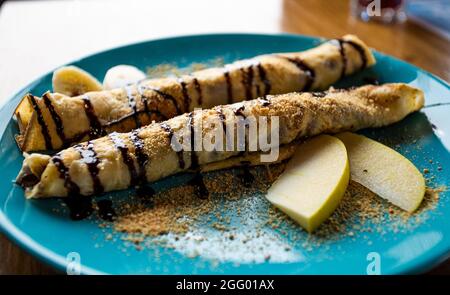 Arranged two crepe pancakes rolls on blue plate with apple and banana slices and chocolate topping. Dessert or sweets on table in restaurant Stock Photo