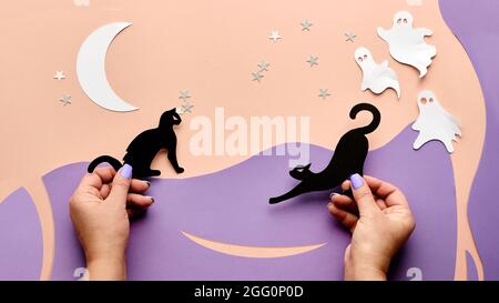Cute Halloween background with copy-space. Hands holding Kawaii black cats and cute white paper ghosts. Flat lay on pink and light purple paper with Stock Photo