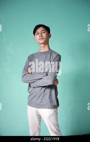 portrait of handsome asian man wearing gray t-shirt with cool pose. skinny man on green background. man folds hands on stomach with serious expression Stock Photo