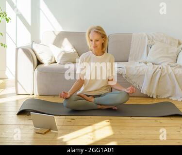Retired woman meditating and practicing yoga while sitting in lotus pose on floor at home Stock Photo