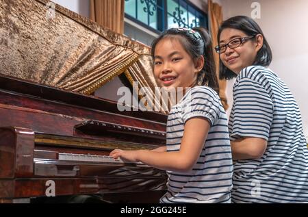 Asian cute child girl practicing pianon lesson white her mother at home, playing upright piano, looking to camera and smile. Stock Photo