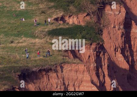 Sidmouth, Devon, UK. 27th Aug 2021.  Members of the public stand dangerously close to the edge of crumbling cliffs whilst watching the RAF Red Arrows perform a colourful display over Sidmouth, Devon. The cliffs at Sidmouth have collapsed in this location around half a dozen times in the past 4-6 weeks. Credit: Ian Williams/Alamy Live News Stock Photo