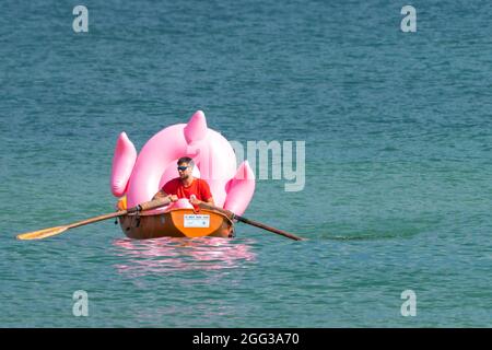 Hel, Poland - 08.01.2021: Beach lifeguard rowing in his orange boat carrying a big pink inflatable flamingo back to the shore on a sunny summer day. Stock Photo