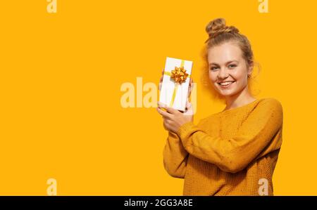 Smiling beautiful blonde young woman holding a gift box isolated over yellow background. Happy birthday, Christmas, Valentine's Day concept. Place for Stock Photo