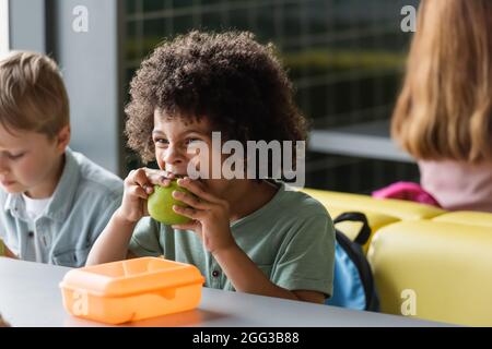 african american schoolboy eating apple near blurred classmates in school canteen Stock Photo