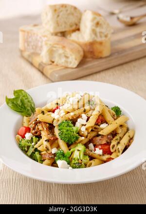 Studio photo of a bowl of penne pasta with broccoli and feta cheese and a cutting board of ciabatta bread slices. Stock Photo