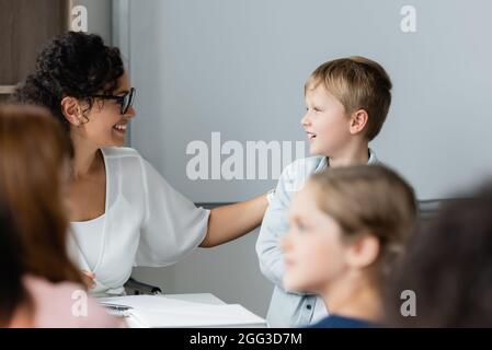 positive african american teacher talking to schoolboy during lesson near blurred pupils Stock Photo